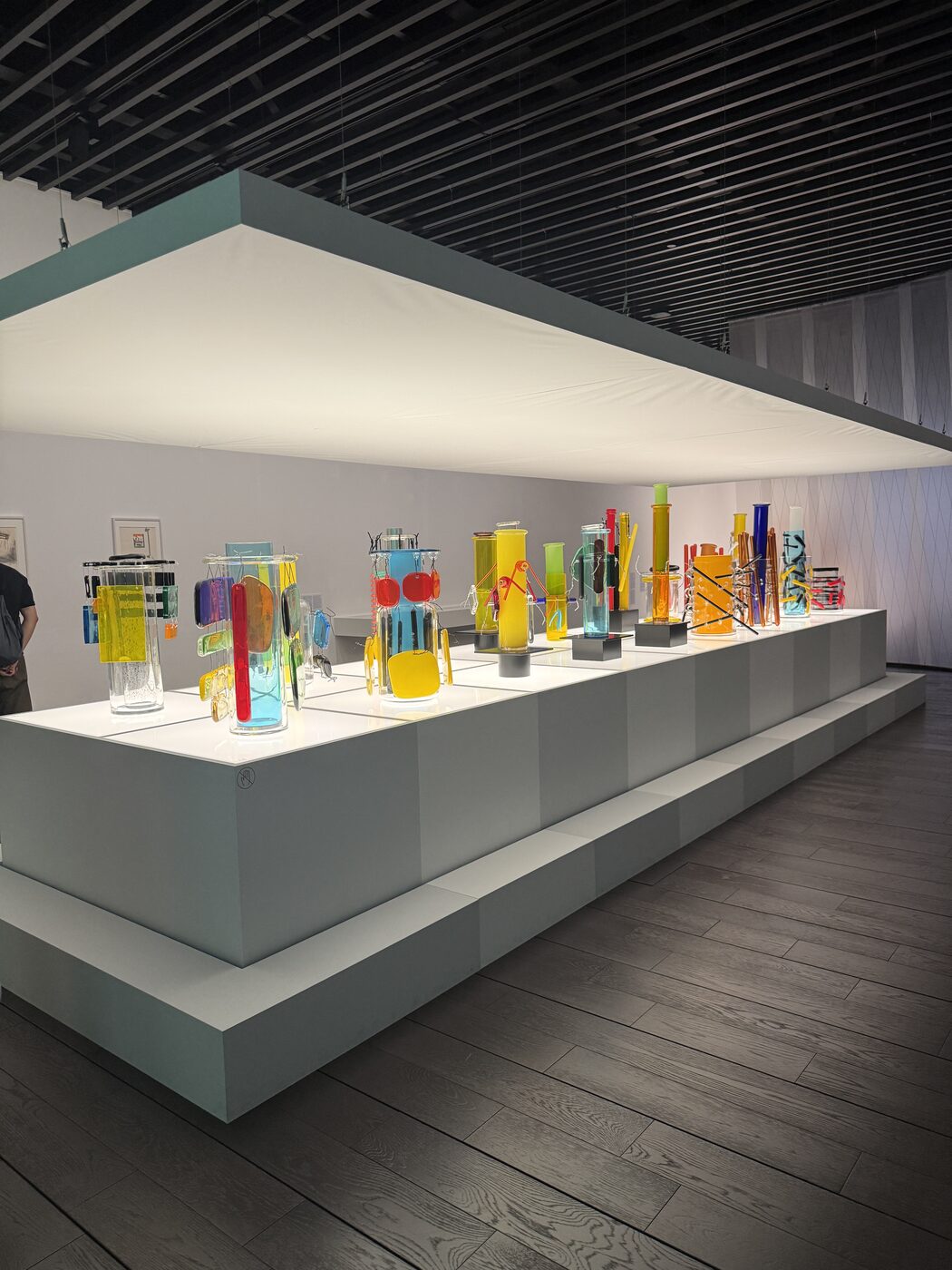 A long lit plinth of colourful Sottsass ceramics and glass objects