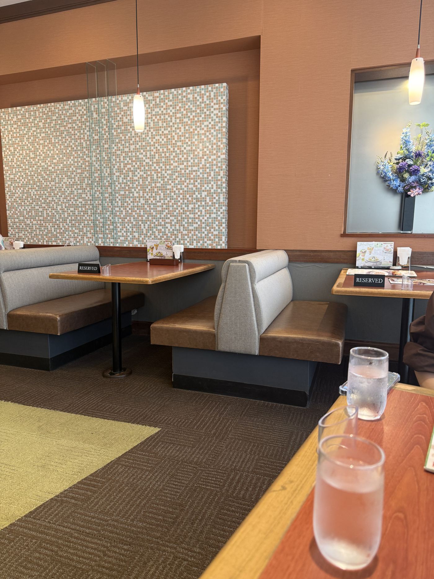 The quiet interior of Royal Host Saitama-Shintoshin: grey and brown booth seating, wooden tables, a patterned wall panel and a vase of blue flowers in a wall niche