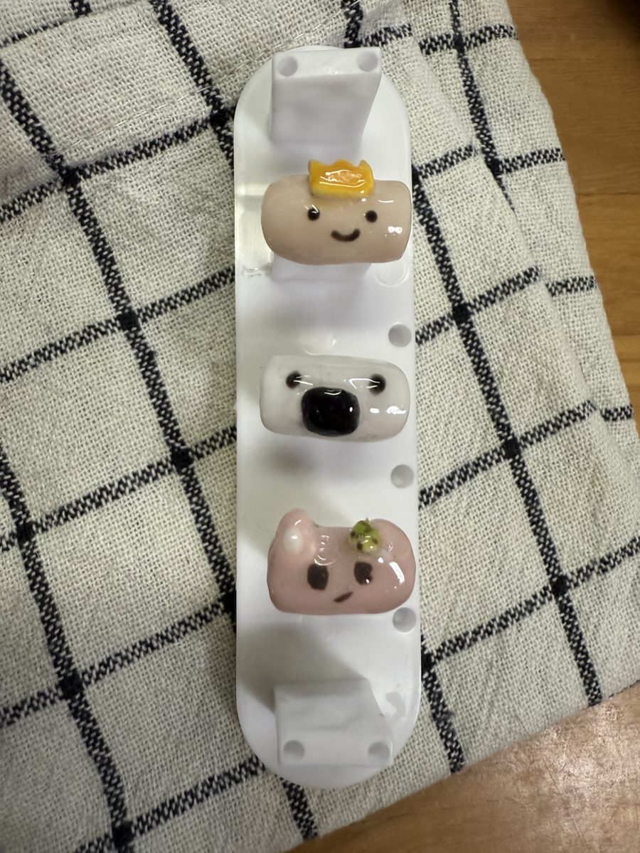 Three small hand-made nail tips on a white holder: one with a yellow crown, one with a black nose, one pink with a green detail